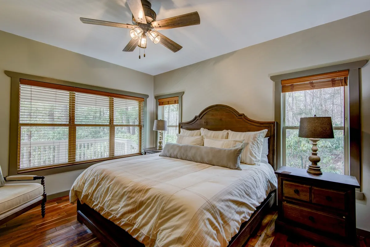 A serene bedroom featuring a plush bed with decorative pillows, wooden nightstands, a ceiling fan with light fixtures, and large windows with blinds that allow natural light to filter in. The space exudes calmness and privacy, ideal for restful nights.