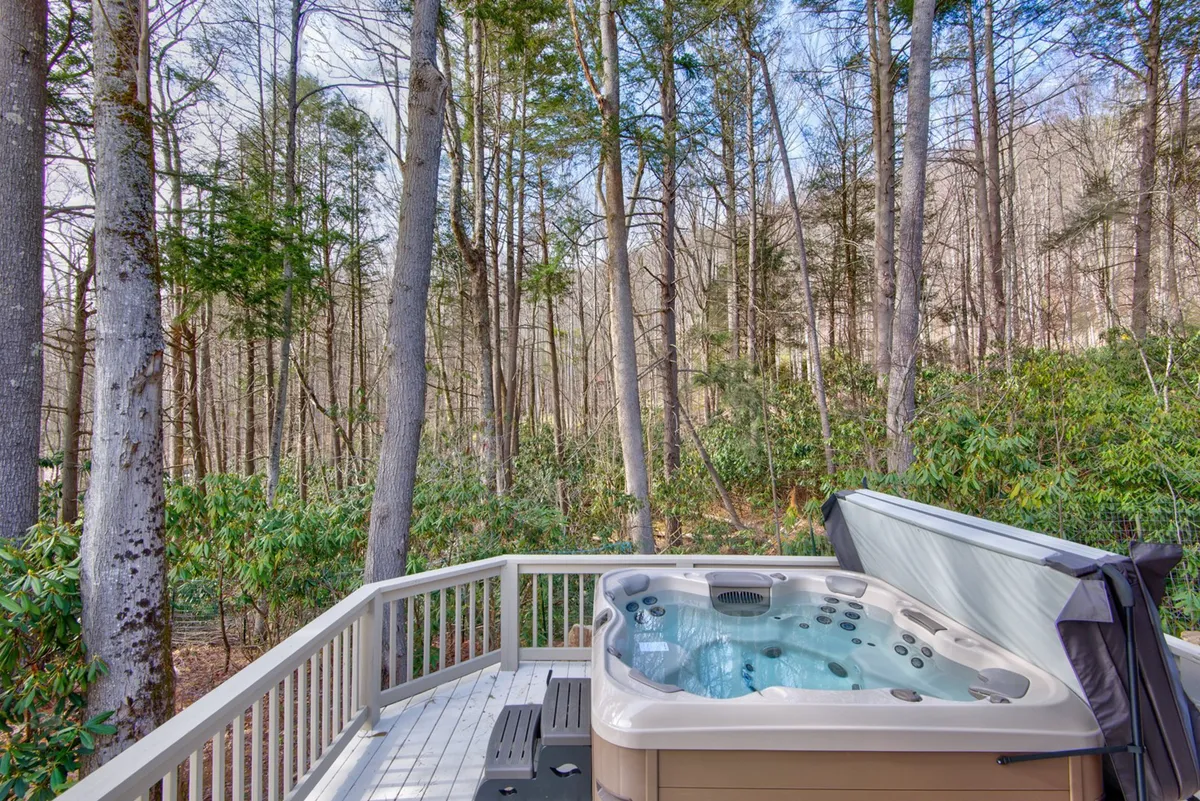 The image captures a serene hot tub area on a wooden deck, surrounded by lush trees and a rustic railing. The sky above hints at the possibility of starlit evenings, and the ambiance invites relaxation while basking in the natural beauty.
