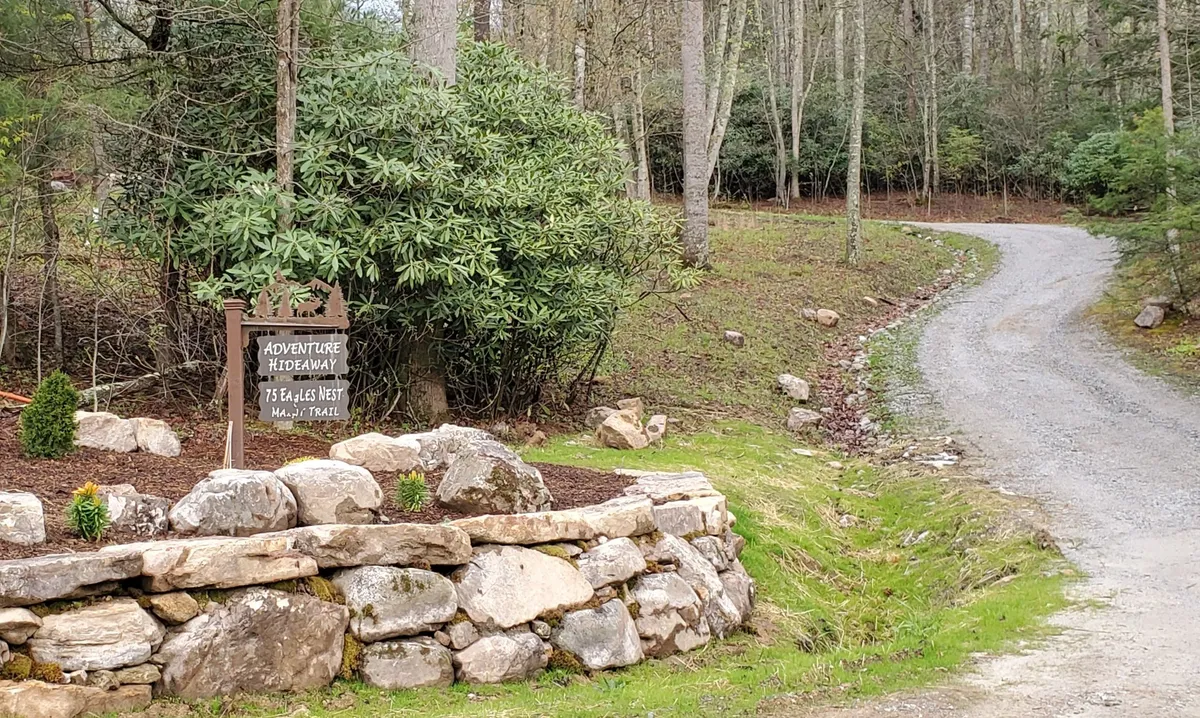 A rustic sign reading 'Adventure Hideaway' marks the property's entry, bordered by a rock wall and lush greenery. The gravel path leads into the woods, suggesting access to hiking and adventure trails right from the doorstep.