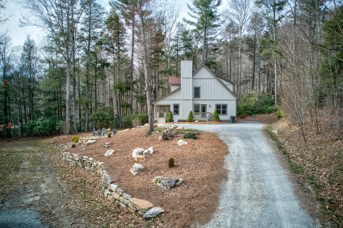 The view highlights the house, the driveway, a landscaped garden, and the wooded backdrop. The open layout and well-maintained grounds suggest plenty of room for dogs to roam, and the surrounding trees reinforce the sense of a mountain escape immersed in nature.
