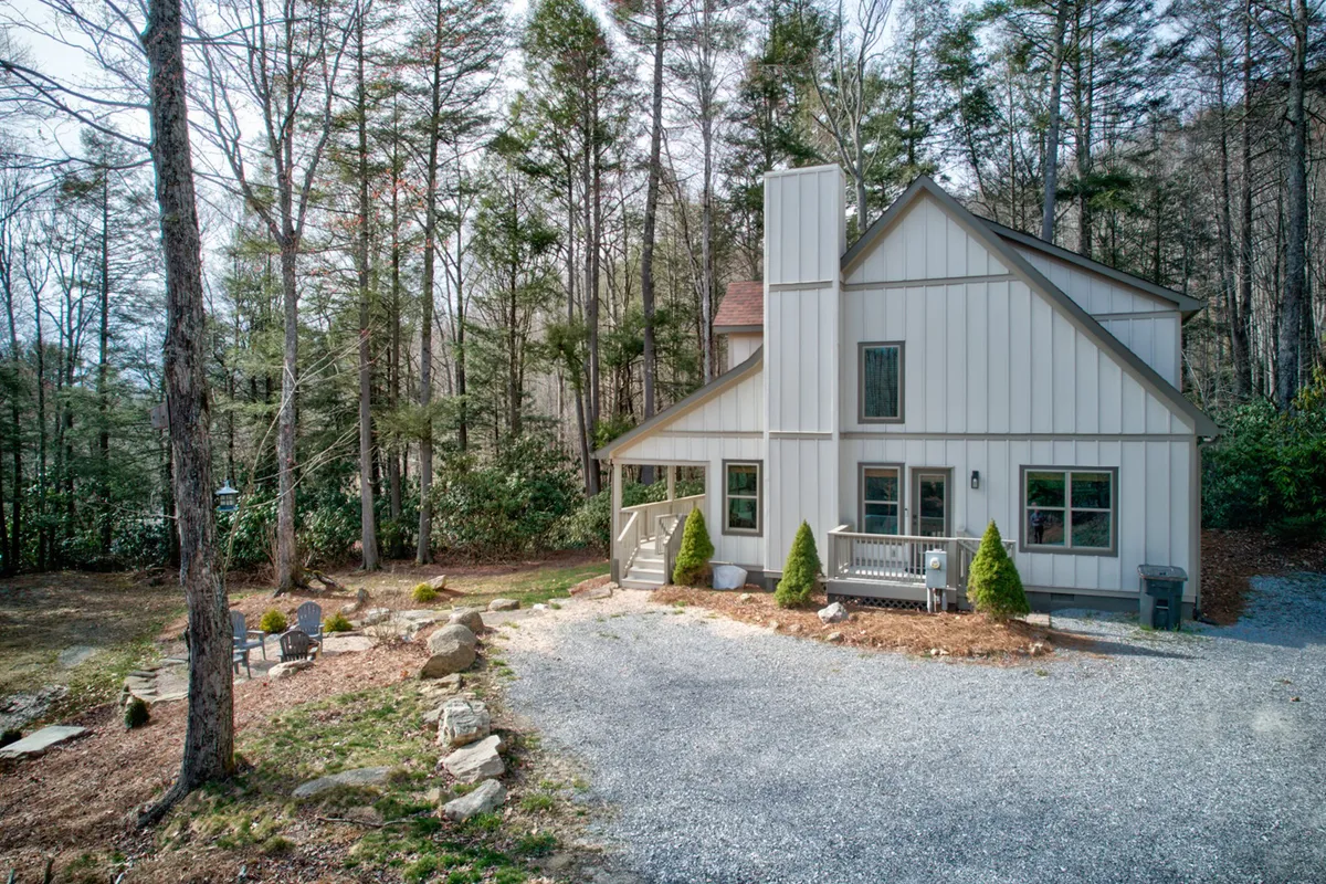 A spacious exterior view shows the home surrounded by trees, with a gravel driveway leading up to a welcoming porch and a cozy outdoor seating area. A fire pit hints at group gatherings, and the lush greenery evokes the setting's connection to nature and trails.