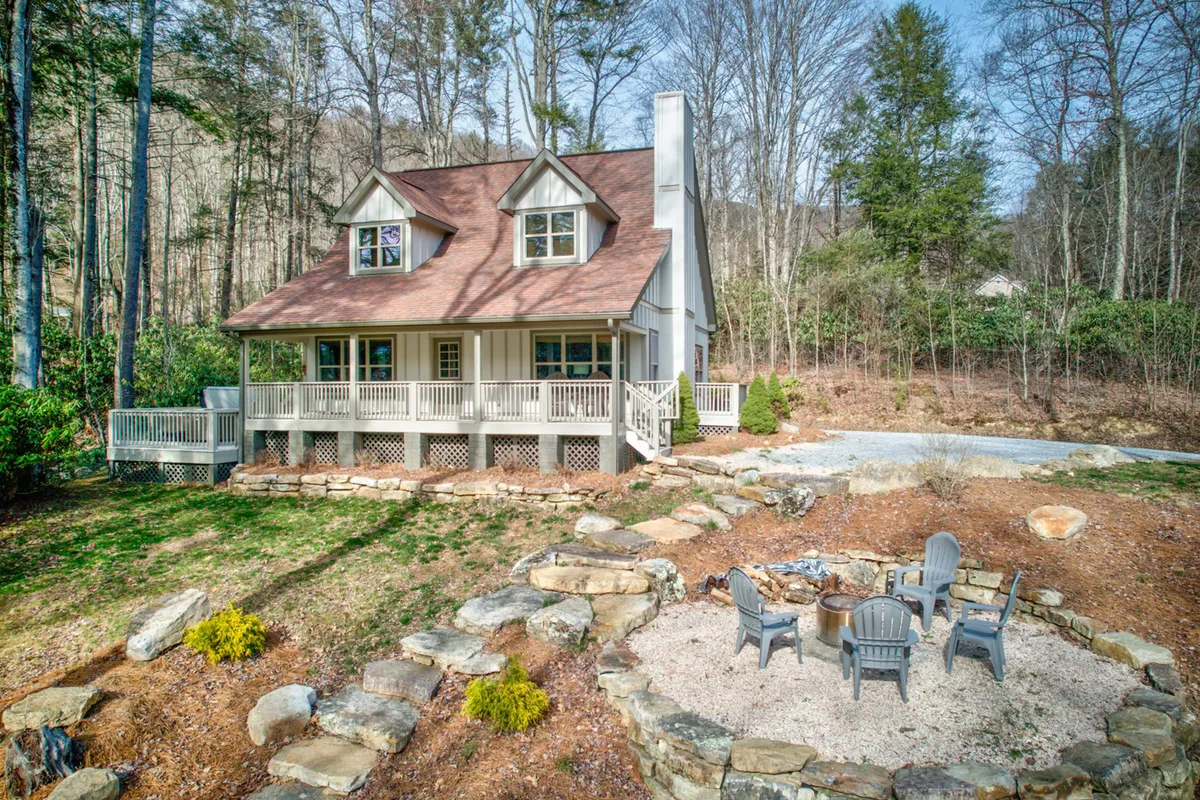 The image shows a charming, modern cabin nestled among tall pines in the scenic mountains of Banner Elk, North Carolina. The inviting outdoor fire pit is set in front of the house, surrounded by comfortable Adirondack chairs, suggesting a perfect spot for gathering with friends and family. The peaceful setting is enhanced by lush trees and a spacious porch overlooking the serene natural landscape, promising relaxation and memorable evenings under the stars.