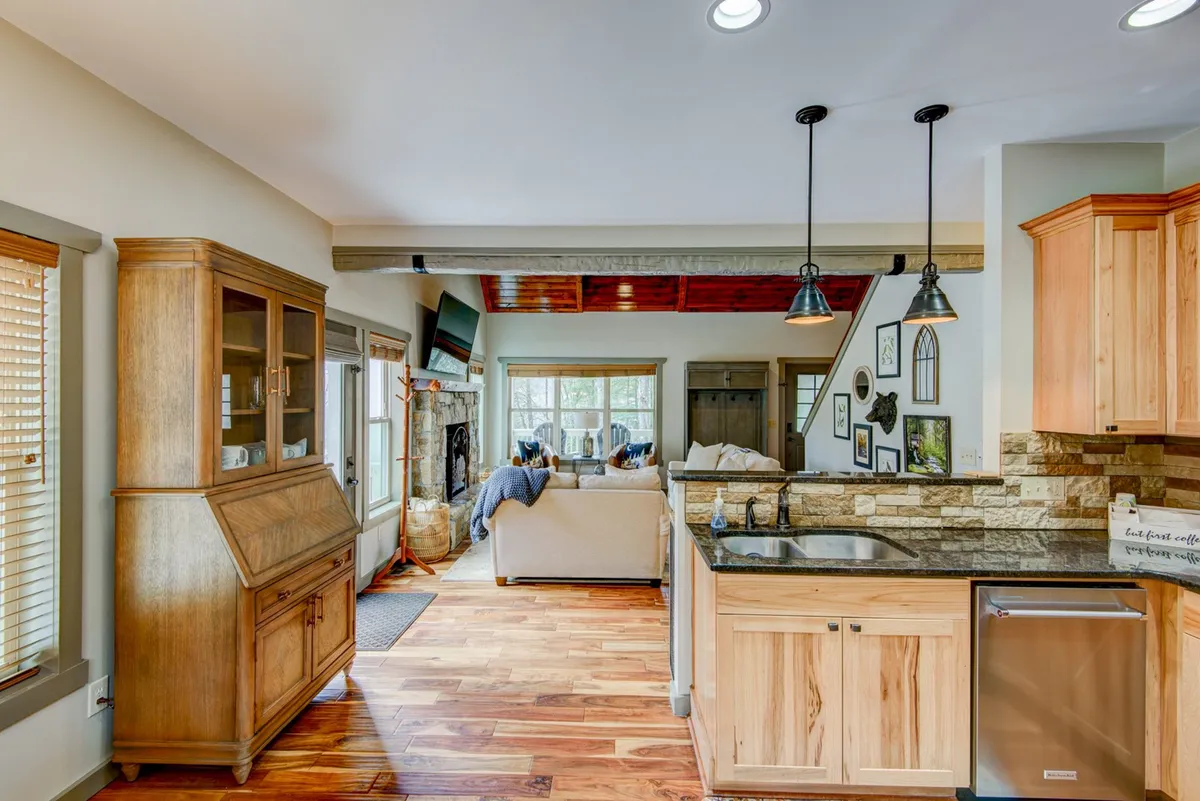 A wide interior view that encompasses both the comfortable living area with a sofa and fireplace and the adjoining kitchen with wooden cabinetry, stone backsplash, and a central island. The layout demonstrates how the spaces flow together for easy, communal living.