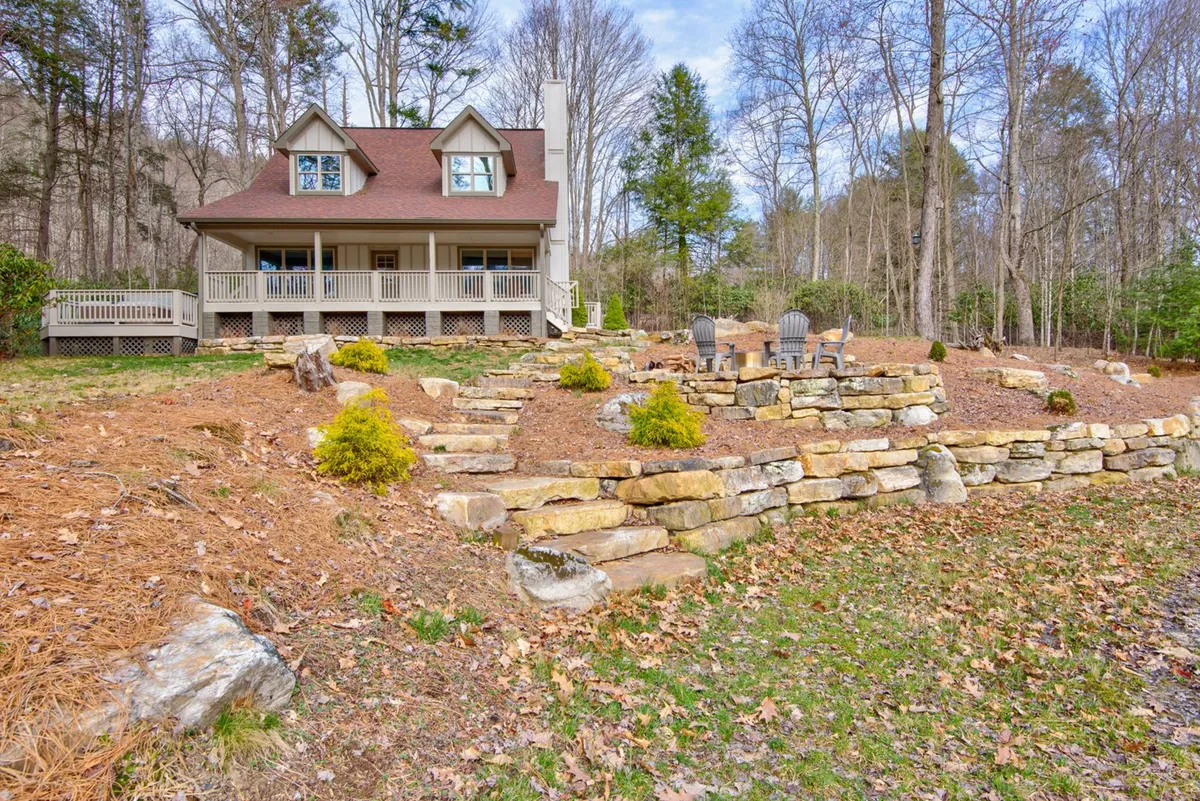 A cozy exterior scene featuring the house with a welcoming porch, stone steps, and a gathering area with chairs set amidst mature trees. The inviting setup suggests both relaxation and togetherness, with the fire pit area hinting at evening s'mores or storytelling.