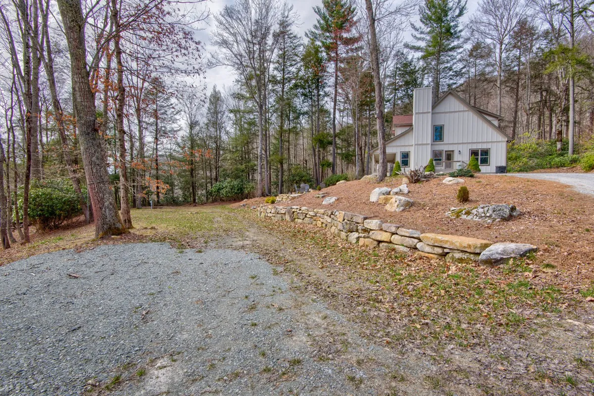 A peaceful scene showing the home's facade, with a stone wall, tree-lined backdrop, and spacious driveway. The setting suggests opportunities for outdoor gatherings, nature walks, and enjoying the calm of the gated community.
