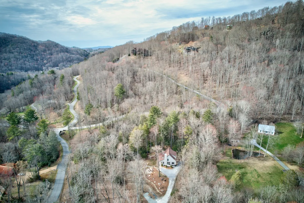 A sweeping view of the Eagles Nest community, showing the central house with a distinctive red roof set among rolling, wooded hills and winding roads. The scene is surrounded by dense forests and open skies, suggesting ample space and natural beauty for outdoor activities and adventures.