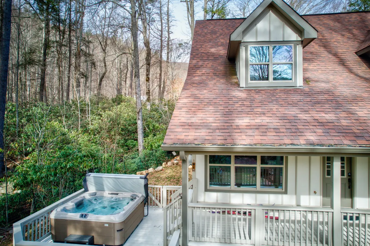 This image highlights a peaceful hot tub set against cottage-style architecture, surrounded by mature trees and natural landscaping. The view hints at the expansive deck and the connection to scenic outdoor spaces, blending both relaxation and the beauty of the environment.