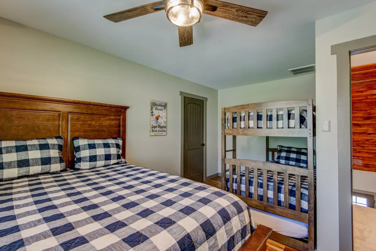 A spacious bedroom with a king-sized bed with checkered bedding, a bunk bed with matching bedding, a wooden ceiling fan, and decorative wall art. The room is inviting and versatile, suitable for families or groups to gather and relax.