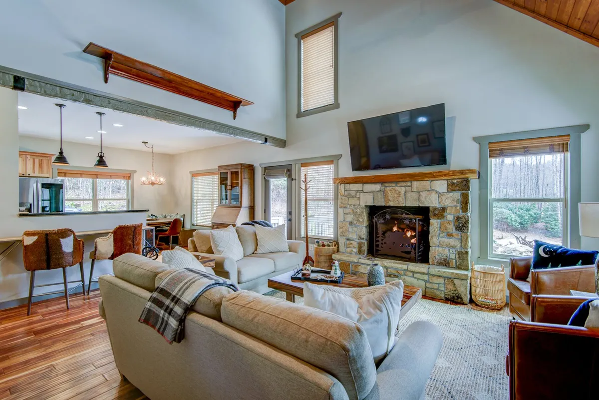 A spacious open-concept living area with a soaring cathedral ceiling, large windows flooding the room with natural light, a central gas fireplace, plush seating, and a view toward the adjacent kitchen bar. The layout highlights both the gathering space and the seamless connection between living and kitchen areas.